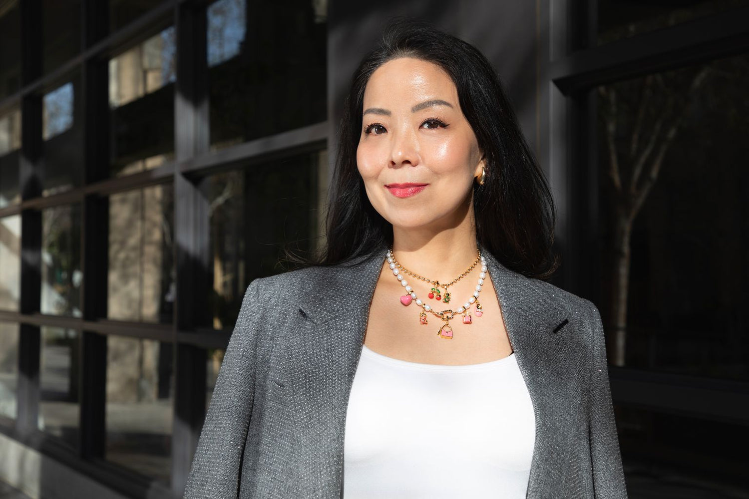 Our founder, Jennifer Jaehwa Yu, in a gray blazer and white top standing outdoors with a building in the background adorned in her jewelry collection: Jyuls by Yu necklace and charms